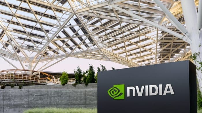 The image shows a large, black sign with the word "NVIDIA" in white letters, prominently displayed in the foreground. The sign is situated in front of a modern building with a white metal roof structure that appears to be made of solar panels. The building's design incorporates a curved, open-air structure, possibly a