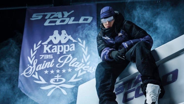 The image shows a person wearing a purple and black jacket, black pants, and a purple beanie, seated on a white platform with a purple banner behind them. The banner displays the text "Stay Cold" and "Kappa 735 Saint Players", along with a graphic of two Kappa Kappa Kappa figures. The person is also wearing white snow