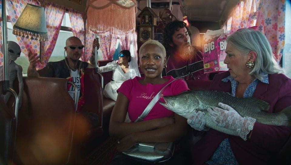 The image shows a group of people seated on a bus, with a large fish being held by one of the passengers. The bus interior is decorated with pink and white floral curtains, and a clock is visible on the wall. The passengers are dressed in a variety of clothing, including a pink shirt with the word "Luck" on it. One pas