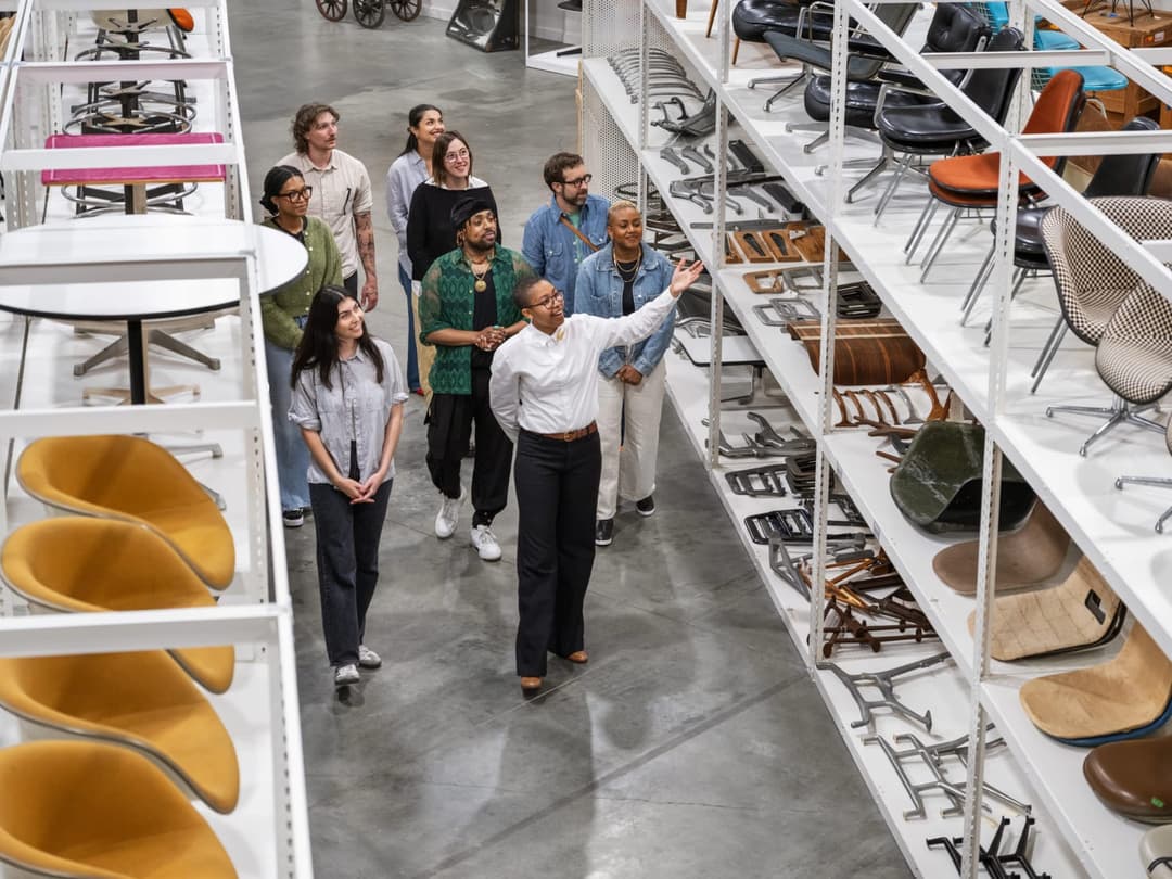 The image shows a group of people standing in a warehouse, examining a variety of chairs and tables. The warehouse has a high ceiling and a gray floor, with white shelves that hold a collection of chairs and tables. The chairs and tables are arranged in a somewhat organized manner, with some tables placed on the left s
