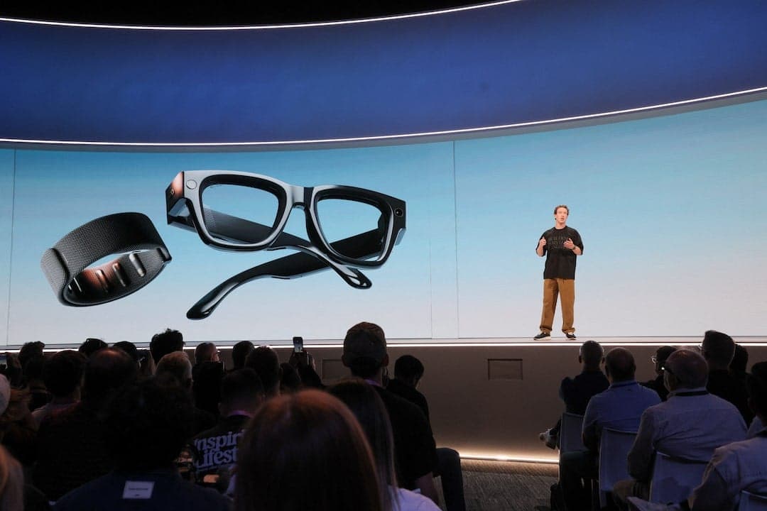 The image captures a presentation taking place on a stage. A man in a black shirt and khaki pants stands at the center of the stage, addressing an audience seated in front of him. The stage is set against a backdrop of a blue wall with a curved design. A large screen behind the speaker displays a pair of black glasses