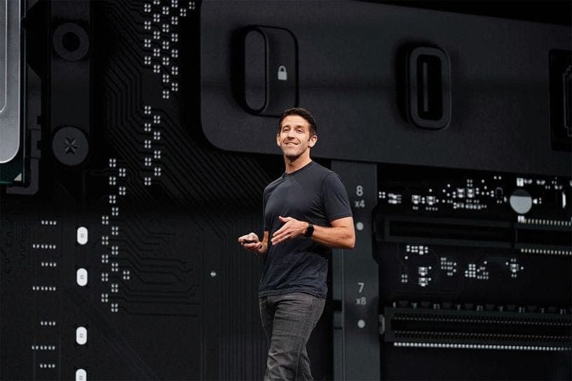 The image shows a man standing in front of a large black computer screen. He is dressed in a black t-shirt and gray pants, and he is gesturing with his hands as if explaining something. The computer screen is adorned with a complex network of white dots and lines, creating a striking contrast against the black backgrou