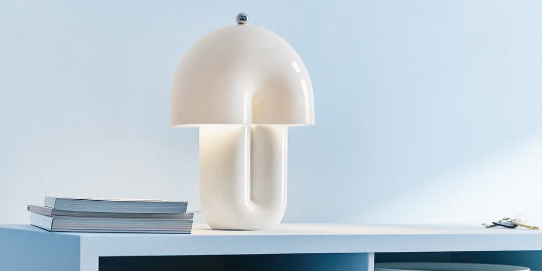 The image depicts a minimalist scene featuring a white lamp with a curved, dome-like shade and a curved base, standing on a white shelf against a light blue wall. The lamp is illuminated, casting a soft glow. To the left of the lamp, there is a stack of books, and to the right, a set of keys are placed on the shelf. Th