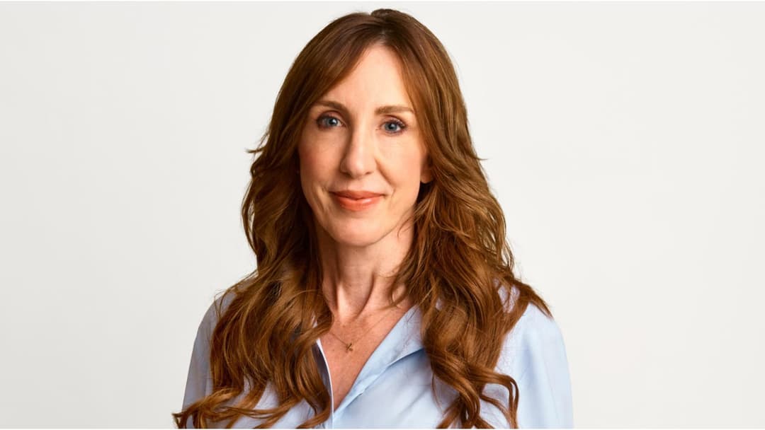 The image shows a woman with long, wavy, light brown hair as the central figure. She is wearing a light blue, collared, button-down shirt and a delicate gold necklace. Her gaze is directed straight at the camera, and she has a warm, friendly smile. The background is a plain white, providing a stark contrast to the woma