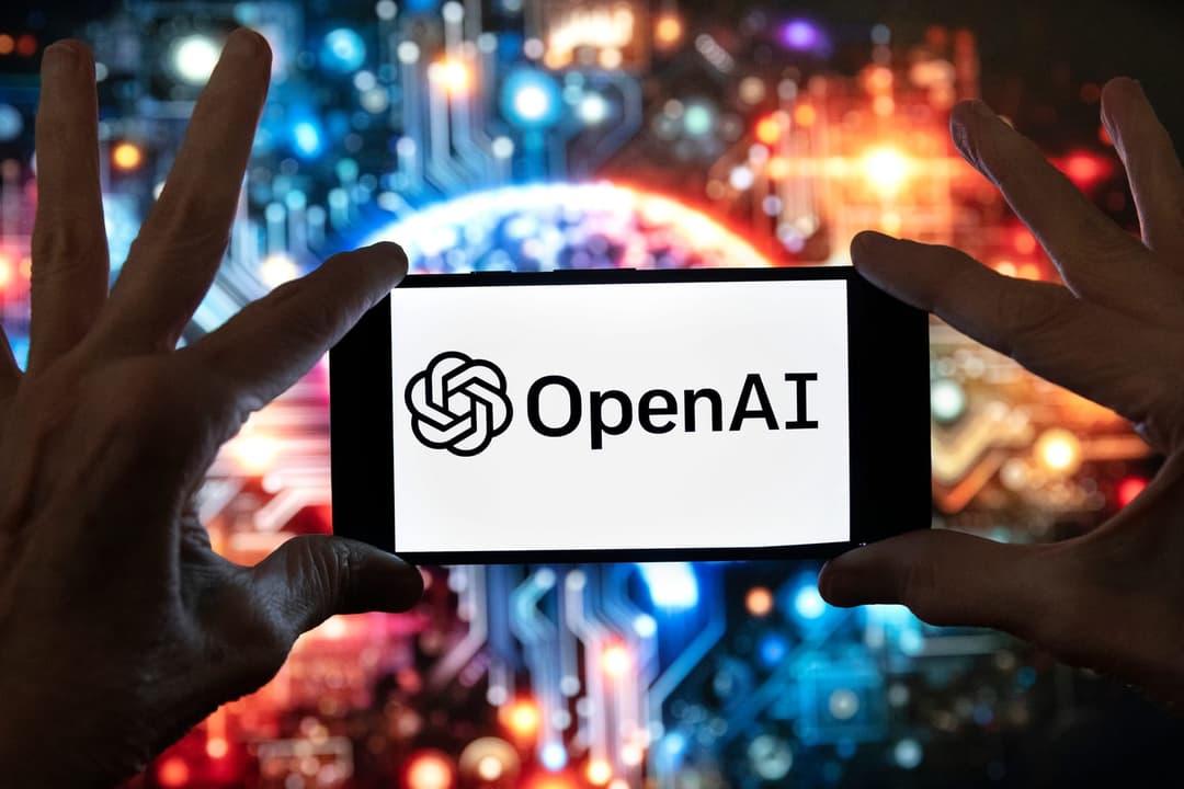 The image shows a pair of hands holding a smartphone in front of a blurred background that appears to be a circuit board. The smartphone's screen displays the OpenAI logo, which is a stylized black and white design. The logo is centered on the screen. The background is out of focus, but it appears to be a mix of blue,