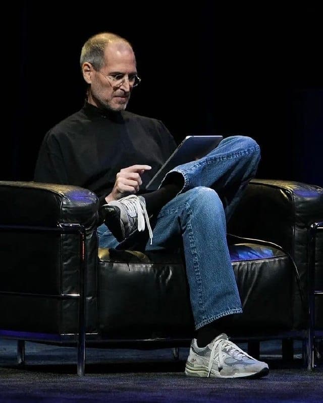 The image shows a man seated in a black leather armchair, engrossed in his work on a tablet. He is dressed casually in a black turtleneck shirt and blue jeans, with white sneakers on his feet. His hair is short and light brown. The man is positioned in the center of the image, with the black leather armchair and the da