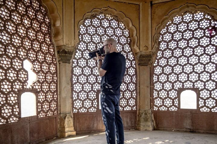 The image shows a man standing in a room with a unique architectural design. He is holding a camera, seemingly capturing the intricate details of the room's design. The room features arched windows with a geometric pattern, and the walls are a light beige or tan color. The man is wearing a dark-colored t-shirt and dark