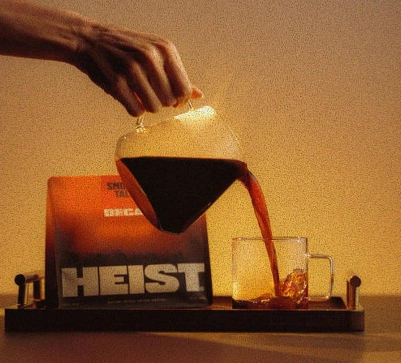 A hand is pouring a dark brown or black coffee from a clear glass pitcher with a handle into a clear glass mug with a handle. The mug is placed on a dark brown wooden tray. The background is a warm, golden-brown color. A black box with the word "HEIST" in white letters is visible behind the tray.