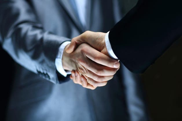 The image shows two individuals in business attire, one in a gray suit and the other in a black suit, shaking hands in a handshake. The gray-suited individual is on the left side of the frame, and the black-suited individual is on the right. The handshake is a firm and respectful gesture, suggesting a professional rela