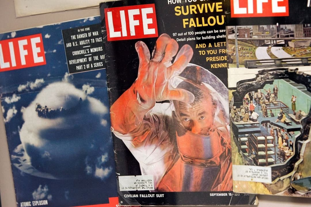 Two LIFE magazine covers are shown, with the center cover featuring a man in an orange plastic fallout suit with one hand raised, headlined "HOW YOU CAN SURVIVE FALLOUT" and dated September 15, 1961. The left cover shows an atomic explosion, the right shows a fallout shelter illustration, and the left cover's text list