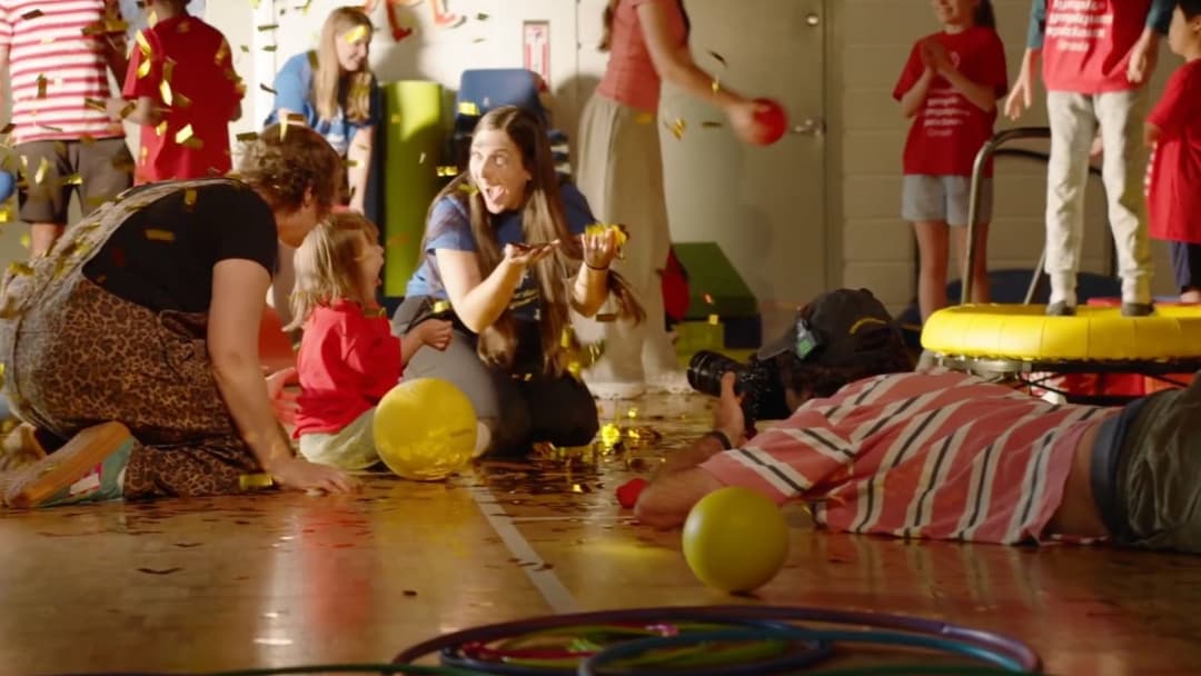 The image captures a lively scene in a gymnasium, where a group of children and adults are engaged in a playful activity. The gymnasium floor is covered in a layer of yellow confetti, adding to the festive atmosphere. A woman in a blue shirt is kneeling on the floor, holding a yellow balloon, while a young girl in a re
