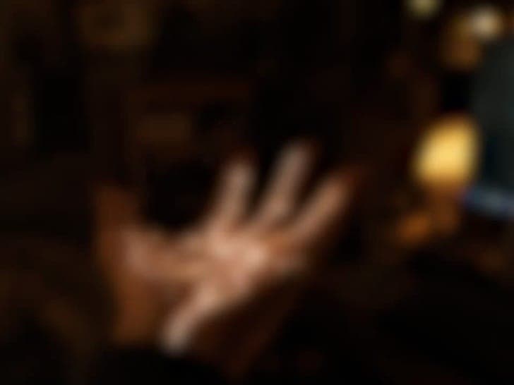The image is a blurry, out-of-focus photograph of a hand reaching towards the right side of the frame. The hand appears to be light-skinned and is reaching towards a light source in the background, which is out of focus and not clearly identifiable. The background is dark and out of focus, with no discernible objects o