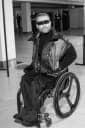 The image shows a man seated in a wheelchair in what appears to be an indoor space, possibly a hallway or corridor. He is wearing a dark-colored turtleneck sweater and a brown vest over it. The man is wearing dark-colored pants and sunglasses. The background is out of focus, but it appears to be a hallway or corridor w