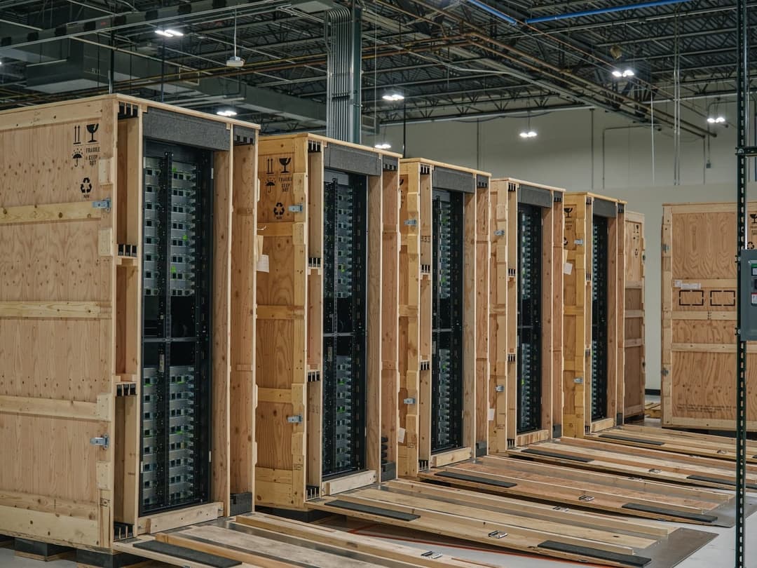 The image shows a warehouse setting with a row of six wooden crates, each containing a server rack. The server racks are primarily black and have a grid-like pattern of green lights. The crates are stacked in a linear fashion, with the first crate on the left and the last on the right. The crates are secured with metal