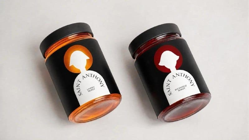 The image shows two jars of Saint Anthony honey, each with a black lid and a black label. The jar on the left is filled with honey that appears to be a light orange or golden color, while the jar on the right contains honey that is a dark reddish-brown color. Both jars feature a white silhouette of a person's head and