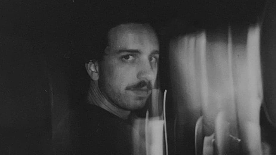 This black and white photograph captures a man with a mustache and goatee, wearing a dark-colored shirt, in a dimly lit room. The man's face is the central focus of the image, with his gaze directed off to the side. The room is dark, with a curtain or fabric visible in the background. The image is slightly blurred, pos