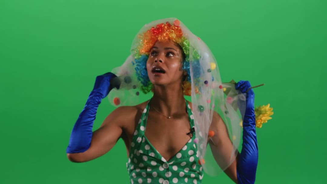 The image shows a person standing against a vibrant green backdrop. The person is wearing a green polka dot dress and a colorful wig with orange, blue, and green hues. They are also wearing blue gloves and holding a small yellow flower in their right hand. The person's face is partially obscured by a sheer veil with co