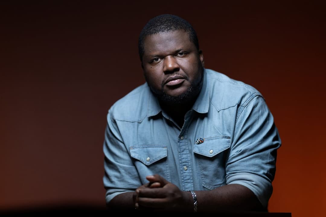 The image shows a man with dark skin and a beard, wearing a light blue denim shirt with a small, possibly gold, pin on the left breast. He is seated at a dark-colored table, with his hands clasped in front of him. The background is a gradient of dark red and orange, creating a warm and intimate atmosphere. The man's ga