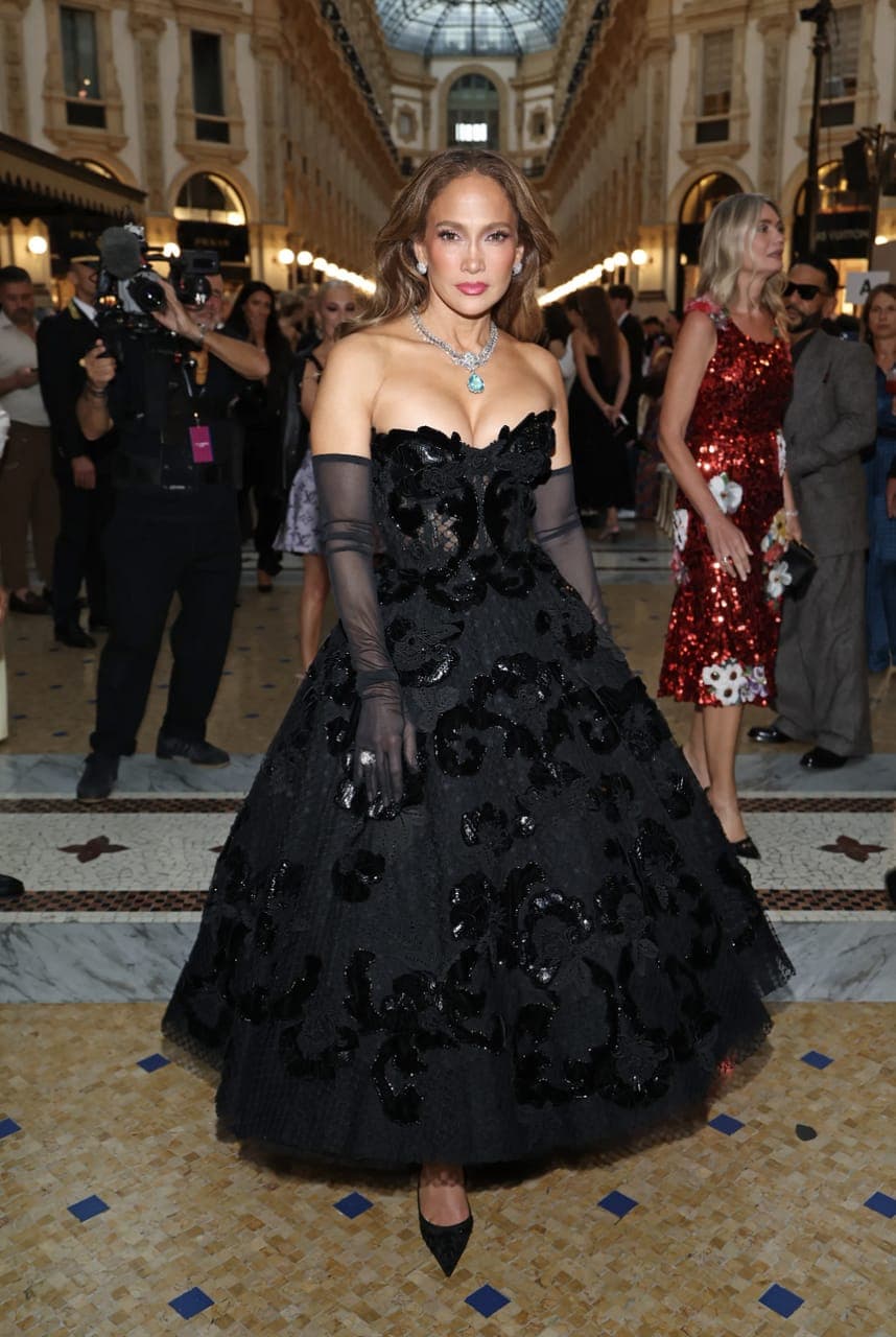 The image shows a woman in a black strapless gown with a floral pattern, elegantly striding down a marble staircase. She is wearing black gloves and a necklace with a turquoise pendant. Her hair is styled in loose waves. The staircase is set against a backdrop of a grand, ornate interior with a glass ceiling and arched