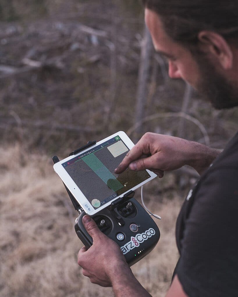 A man stands in a field of dry, light brown grass, engrossed in operating a drone controller. He holds the controller in his left hand and the tablet in his right hand, pointing at the screen with his right hand. The drone controller is black with a white screen displaying a map or terrain information. The man is weari