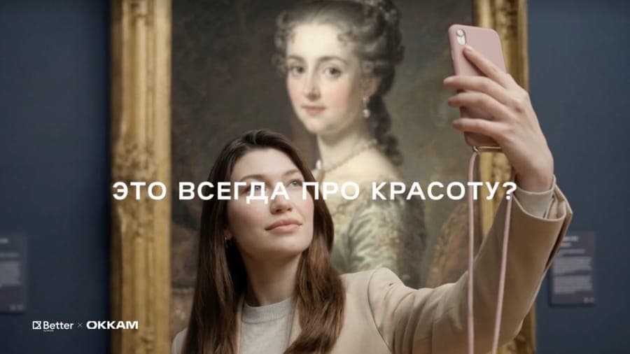 The image shows a young woman with long brown hair, wearing a beige jacket, standing in front of a large, ornate painting in a museum. She is holding a pink phone in her right hand and appears to be taking a selfie. The painting depicts a woman in a period dress, possibly from the 18th or 19th century, and is framed in