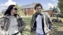 The image shows two individuals standing in a grassy area in front of a large, red brick house with a gray roof. The person on the left is wearing a gray jacket and sunglasses, while the person on the right is wearing a gray jacket and a green scarf. The house has a dark brown or black door and windows. The background