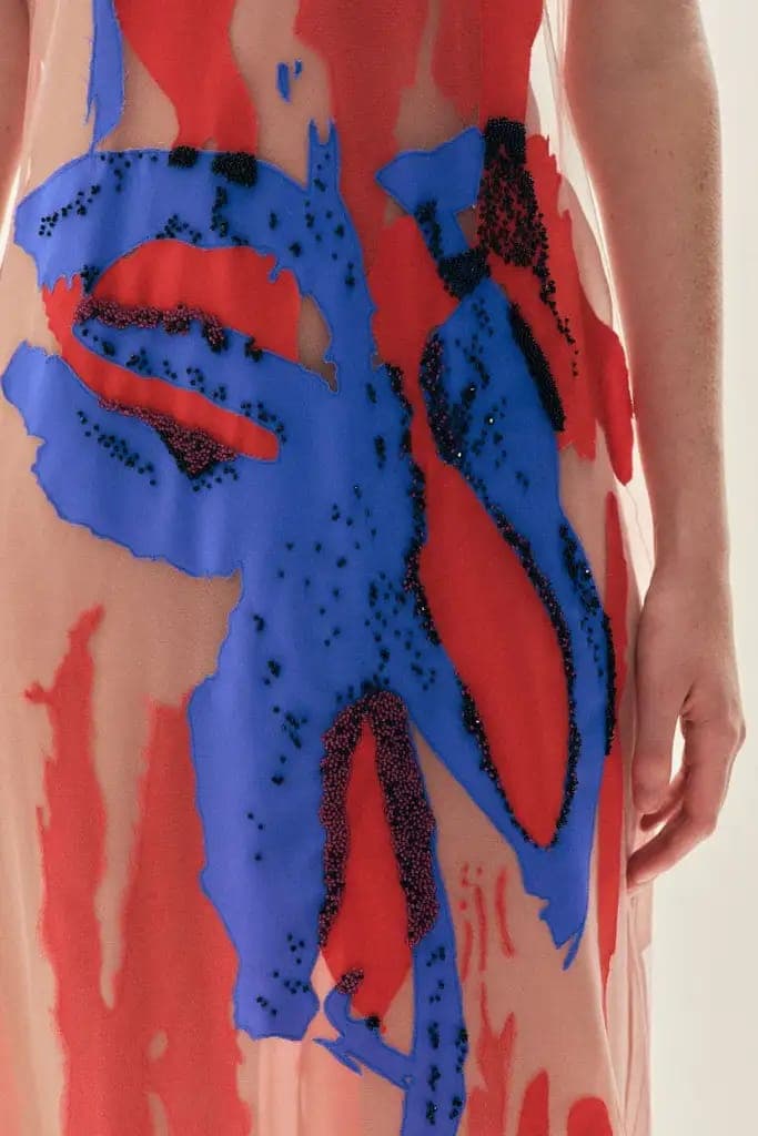 The image shows a close-up view of a person's torso, focusing on a vibrant dress with a unique abstract design. The dress features a blend of red and blue colors, with the blue fabric appearing to be a sheer or semi-sheer material. The design is abstract and incorporates splashes of red and blue, with black dots scatte