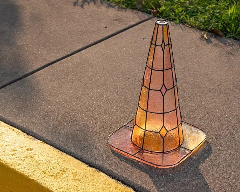 The image shows a traffic cone with a stained glass design, featuring a mix of orange, pink, and yellow hues, resting on a concrete sidewalk. The cone has a black metal frame and a pink base. The sidewalk is bordered by a yellow curb, and a patch of grass is visible in the background. The cone casts a shadow on the sid