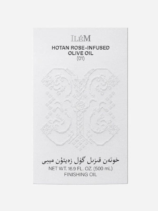 The image shows a rectangular white label with a textured surface, possibly embossed, featuring a floral design. The label is bordered in black and has a small, dark gray/black text at the top and bottom. The text at the top reads "ILÉM" and the text at the bottom reads "HOTAN ROSE-INFUSED OLIVE OIL (01)". The label al