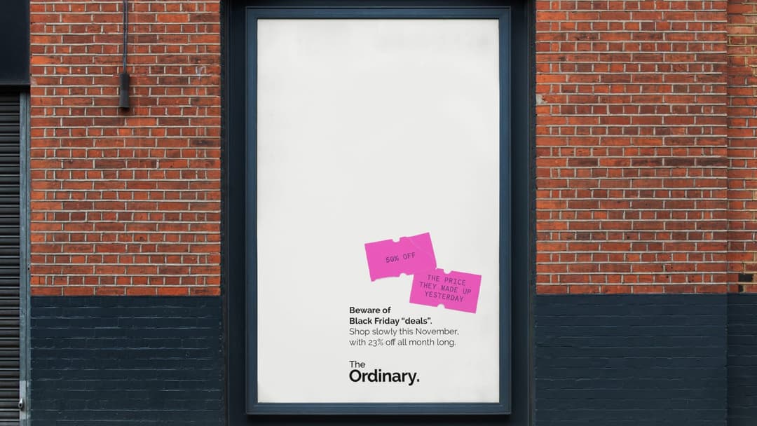 The image shows a brick building with a black frame around a white poster. The poster features a pink banner with the text "50% OFF" and "THE PRICE U PADLASTY" in bold black letters. It also includes a small pink graphic of a price tag. The poster is positioned in front of a dark gray or black wall. The text on the pos