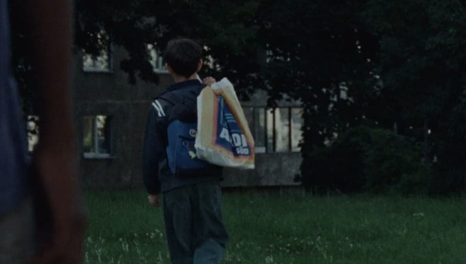 The image shows a young boy walking away from the camera in a grassy area. He is wearing a dark blue jacket and dark-colored pants, and is carrying a white bag with blue and yellow accents. The bag has the word "ADI" printed on it. The boy is walking towards a dilapidated, two-story house with a gray exterior and sever