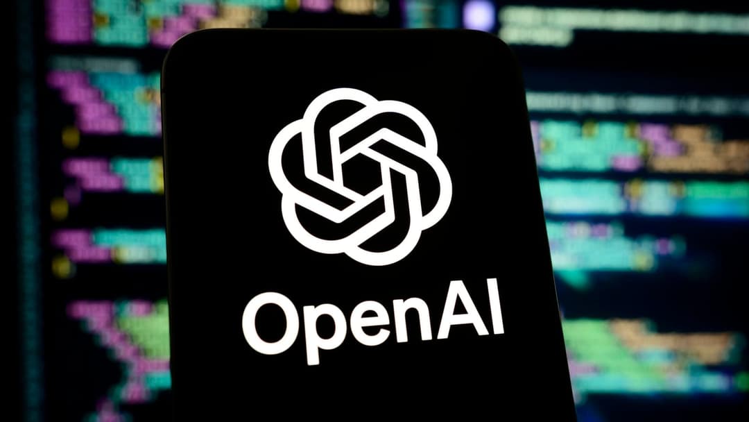 The image shows a black smartphone with a white logo prominently displayed on the screen. The logo is a stylized flower or knot design, and the text "OpenAI" is written in white below it. The smartphone is positioned in the foreground, with a blurred background that appears to be a computer screen displaying lines of c