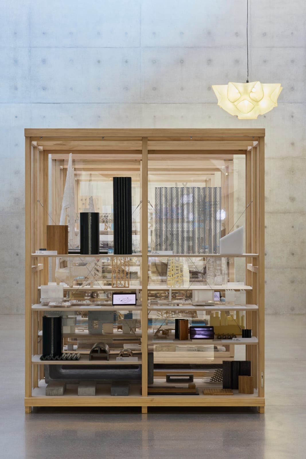 The image shows a wooden display case, possibly made of plywood, with a light brown hue, situated in a room with a concrete wall. The case is divided into two sections, each containing a collection of architectural models and objects. The left section features a variety of models, including a tall black cylindrical mod