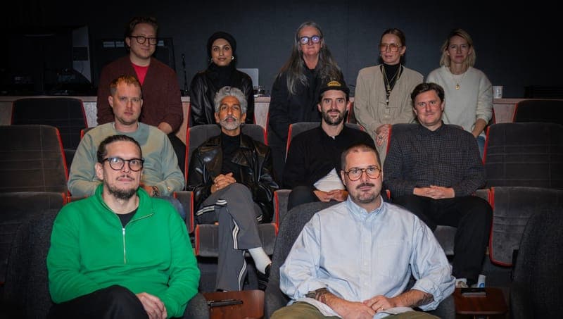A group portrait of nine people seated in rows of dark theater seats, with a man in a green sweater and glasses in front and a man in a light blue shirt at the bottom right. The image has no visible text or logos.