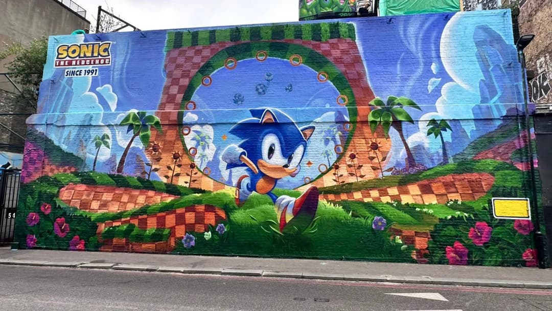 The image depicts a vibrant mural painted on a brick wall, featuring the iconic character Sonic the Hedgehog in a dynamic pose. Sonic is depicted in a blue and white color scheme, running towards a large green and orange checkered archway that serves as a gateway to a fantastical landscape. The landscape includes palm