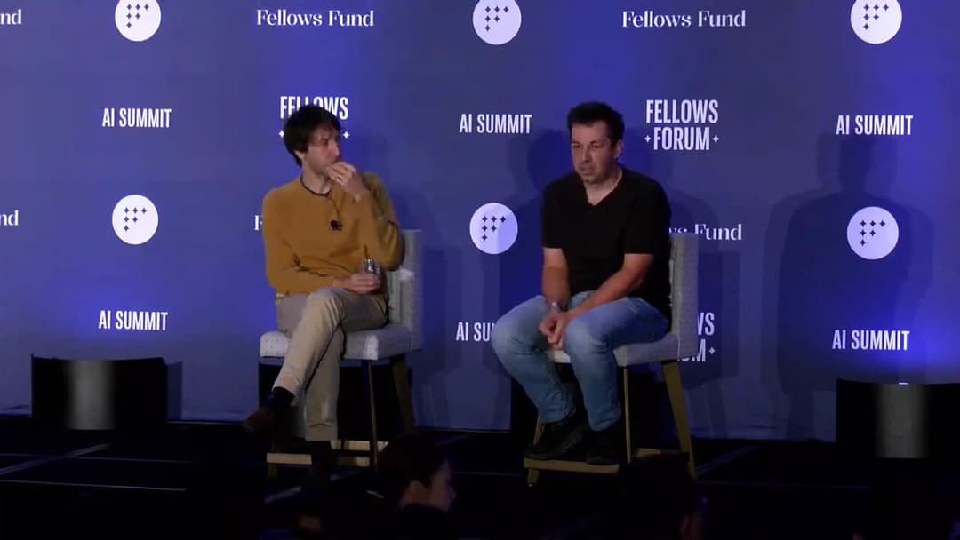 Two men sit in gray upholstered chairs on a stage in front of a blue backdrop with the repeated text "Fellows Fund", "AI SUMMIT", and "FELLOWS FORUM" and a white circular logo. The man on the left wears a mustard sweater and beige pants, and the man on the right wears a black t-shirt and jeans.