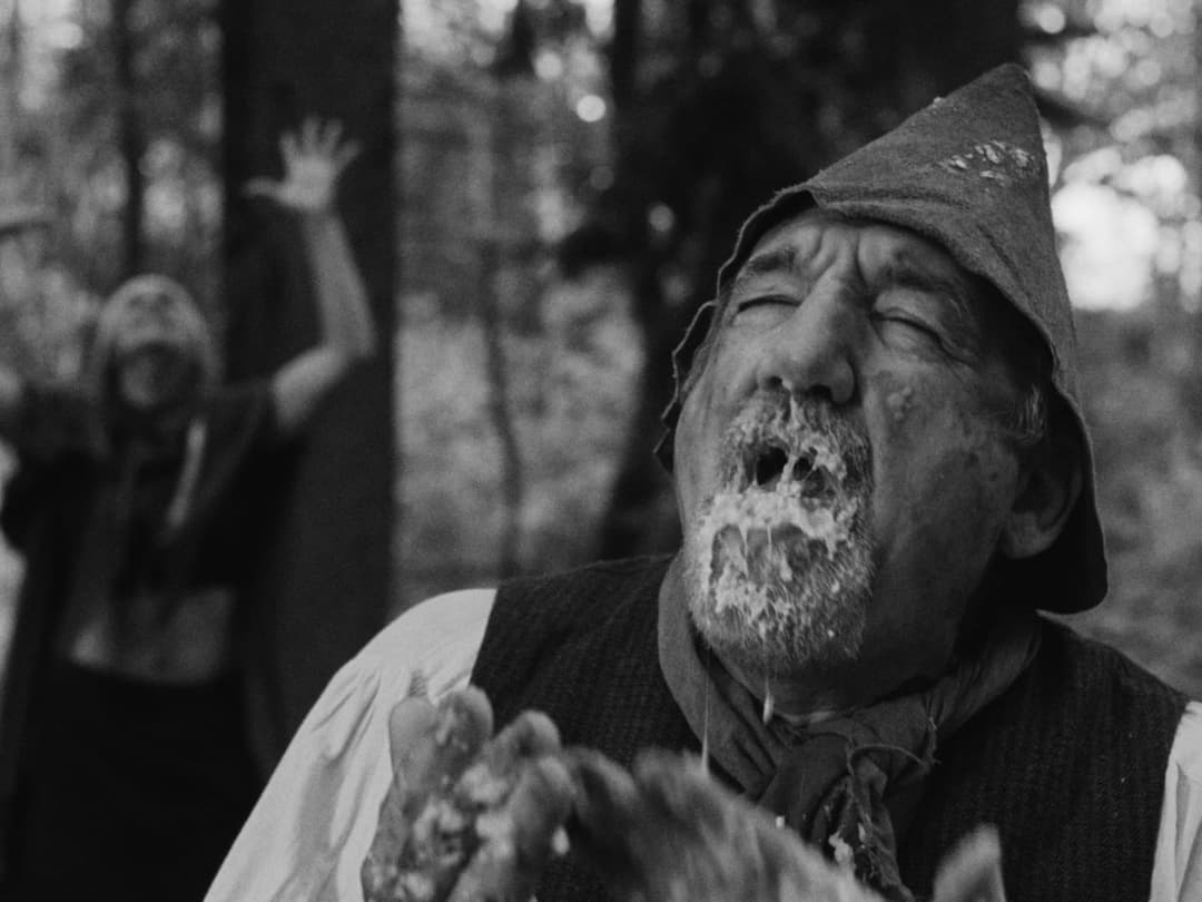 This black and white photograph captures a man in a hat and a dark vest, with a beard and mustache, in a wooded area. He is holding a piece of food, possibly a piece of bread or pastry, and appears to be eating it. In the background, another person is visible, with their arms raised, possibly in a gesture of surprise o