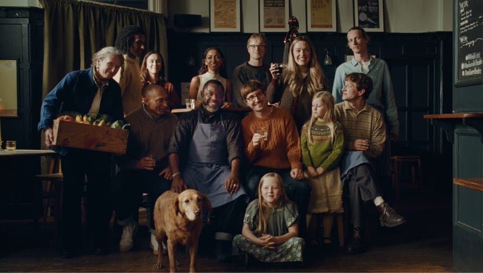 A group of people, including adults and children, are gathered in a dimly lit room, possibly a pub or restaurant. The room has dark wood paneling and a dark green or black wall. A wooden table is visible in the background, and a dark-colored object, possibly a guitar, is positioned near the center of the group. The ind