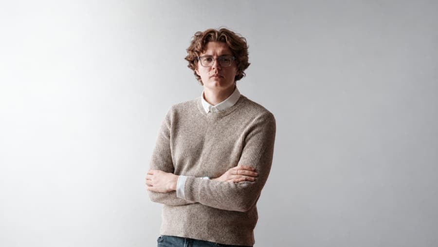 The image shows a young man standing against a plain white wall. He is wearing a beige sweater over a white collared shirt and blue jeans. His arms are crossed over his chest, and he is wearing glasses. His facial expression appears neutral or slightly serious. The simplicity of the background draws attention to the ma