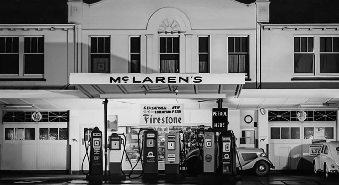 The image depicts a vintage gas station, likely located in the United Kingdom, as indicated by the "McLaren's" sign. The station is housed in a white building with a large arched entrance and a sign that reads "McLaren's" above it. The gas station features a canopy over the entrance, and a sign above the entrance reads
