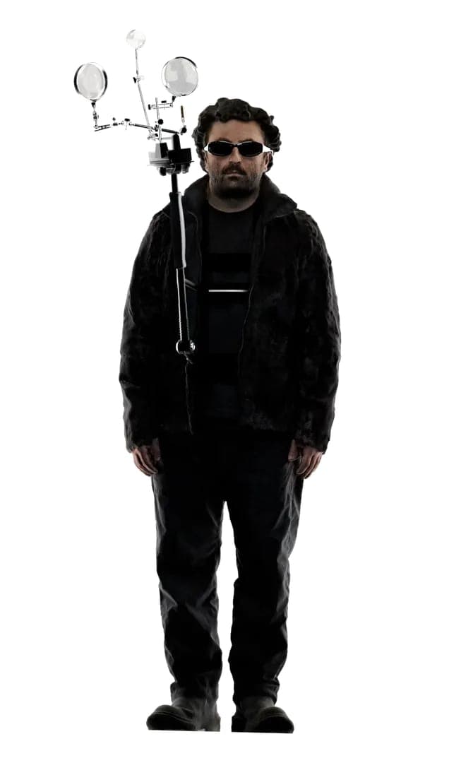 The image shows a man standing against a black background. He is wearing a black jacket, a black shirt with a white stripe, and black pants. He is also wearing sunglasses and has dark hair. He is holding a black pole with a white light fixture attached to it. The light fixture has two circular lights on top and a verti