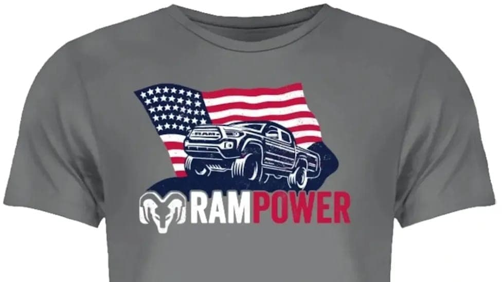 The image shows a gray t-shirt with a graphic design featuring a black and white illustration of a Ram pickup truck positioned in front of a stylized American flag. The truck is depicted in a side profile view, facing towards the right side of the image. The flag behind the truck is a standard American flag with red an