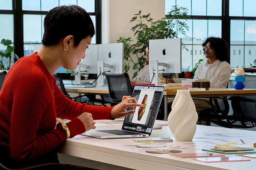 A woman in a red ribbed sweater points at a MacBook Pro screen showing a 3D vase-like shape in a bright office, with iMac computers displaying Apple logos in the background. The image is an Apple-style product photo.