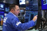 The image shows a man in a blue suit, possibly a stockbroker, seated at a desk in a room filled with multiple computer monitors. He is intently focused on the screen in front of him, which displays a complex array of data and graphs. The room is filled with other computer monitors and screens, suggesting a busy work en