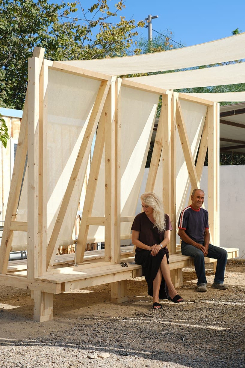 The image shows a wooden structure, possibly a shelter or pavilion, constructed from light-colored wood. It features a white canopy supported by wooden beams and is situated in a gravel area. Two individuals are seated on a wooden bench beneath the canopy. One person is a woman with long blonde hair wearing a dark-colo