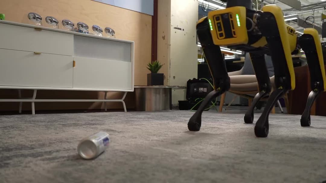 The image shows a robot with a yellow body and black legs standing on a gray carpeted floor. The robot's head is black and has a yellow/gold colored section. It is positioned in the center of the image, facing towards the right side. In the background, a white dresser with a glass front and a small potted plant are vis