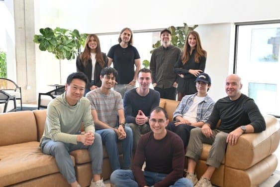 About ten people pose together in a bright indoor space with fiddle-leaf plants, several of them seated on tan leather sofas. No visible text or brand.