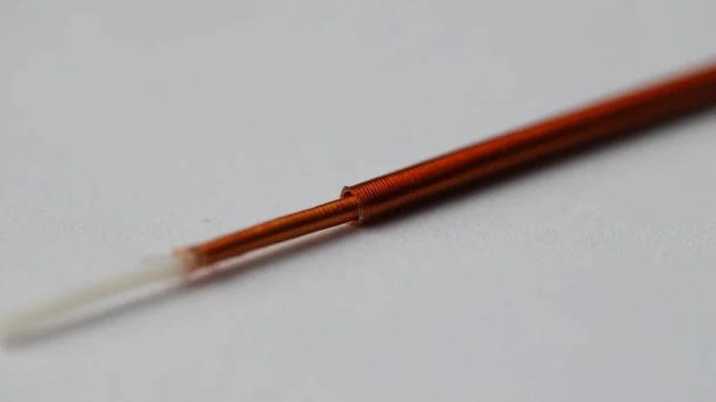 The image shows a close-up view of a single, thin, copper-colored wire. The wire is slightly bent and has a white or off-white end, possibly a connector or part of the wire's structure. The wire is positioned diagonally across the frame, with its tip pointing towards the top left corner. The wire is set against a stark