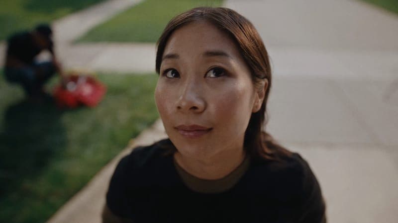 The image shows a young woman with brown hair, wearing a black shirt, standing on a sidewalk. She is looking off to the side with a thoughtful expression. The background is out of focus, but shows a grassy area and a paved path. A person is partially visible in the background, possibly crouching or kneeling. The woman'