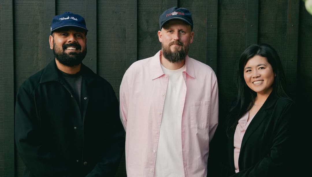 The image shows three individuals standing in front of a dark brown wooden wall. The person on the left is wearing a black jacket and a blue baseball cap with the words "Uncle Pat" written on it. The person in the middle is wearing a pink shirt and a dark blue baseball cap with a logo. The person on the right is wearin